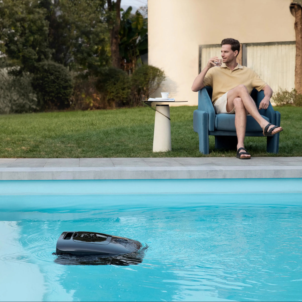 Robotic pool cleaner on the water surface of a pool while a man relaxes in a lawn chair beside it