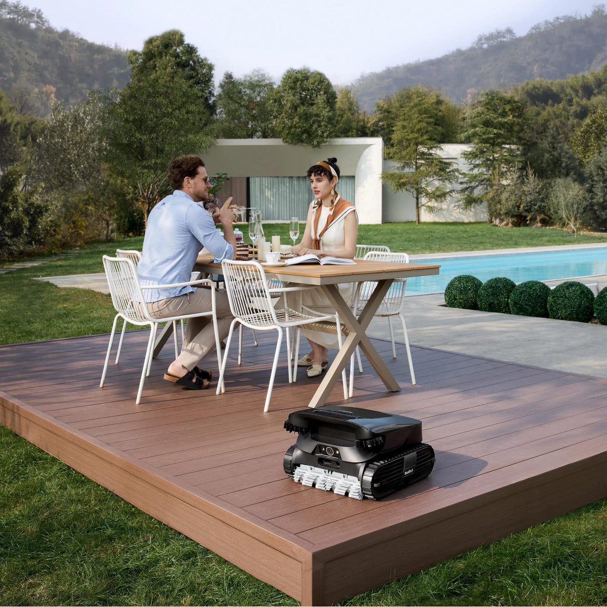 Robotic pool cleaner with a tracked body resting on a wooden deck beside a pool while two people dine nearby